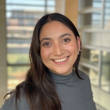 Adelis Cruz smiling in a grey turtleneck sweater on an window background. 