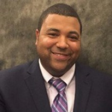 Hector Diaz smiling in a lavender shirt, purple tie, and black blazer on a gray background. 