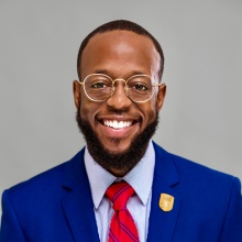 Terrance Lewis smiling in a blue blazer and red tie on a gray background. 
