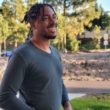 Akil Fletcher smiling with dark shirt in an outdoor background with trees. 