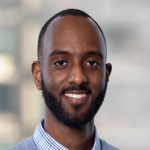 Hamdi Adam smiling in a checkered shirt and gray sweater on an outside background. 