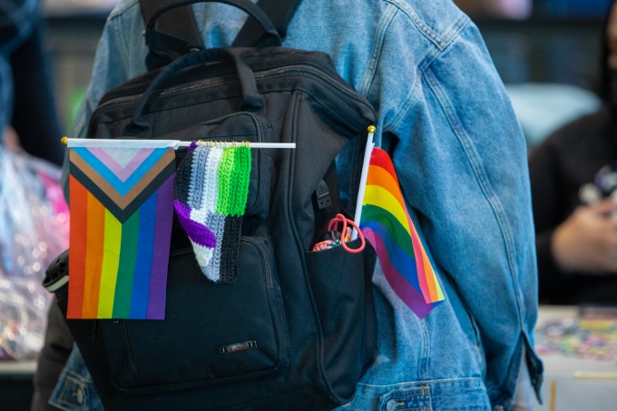 Image of a student wearing a backpack containing a rainbow flag, an LGBTQ+ progress pride flag, and a knitted gay men's pride flag. 