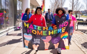 Image of members of the Intercultural and Diversity Center leading the Pride March. They are holding a banner which says "Hate Has No Home Here" with multicolored letters. The banner also includes a hand holding up a transgender pride colored heart, a hand holding up a heart that says "Black Lives Matter," a hand holding up a heart with the American flag, a hand holding up a purple heart with the peace symbol, and a hand holding up a rainbow colored heart. 