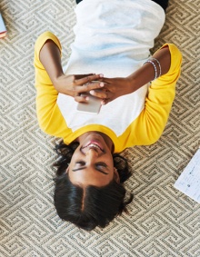 female student in a white and yellow shirt laying on the floor on her back looking at her cell phone with books and notebooks on the floor surrounding her. 