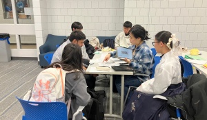 A group of students sitting on one side of a table learning the Korean alphabet from Korean students wearing traditional Korean clothing.