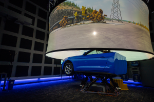 Passenger vehicle mounted on a motion platform inside a driving simulation lab, positioned beneath a large curved projection screen displaying a roadway with trees and utility structures. 