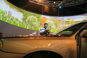 A man stands and appears to take a photo with his phone of a car. a virtual street is displayed on the walls around the man. 