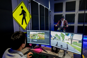 Indoor laboratory with motion‑tracking equipment where one person rides a stationary bicycle while wearing a headset, and another person monitors the simulation on two computer screens displaying a virtual neighborhood street scene; a pedestrian crossing sign is mounted on the wall nearby. 