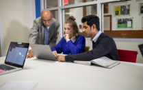 Three individuals sitting at a table, looking at a laptop screen. 