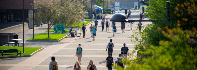 About 20 students walk along the academic spine in front of the Student Union on a warm summer day. The trees and grass are bright green and most students are dressed in summer apparel. 