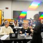 Image of faculty in a training workshop. The facilitator is speaking to two individuals while others are in the background having small group discussions or taking notes. 