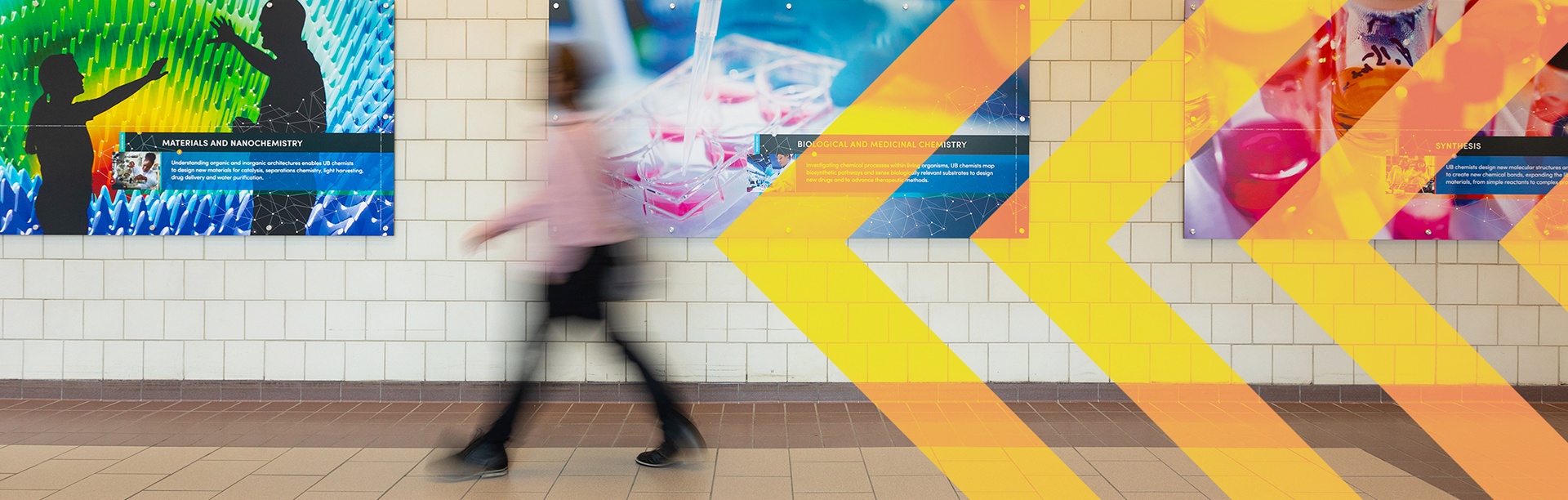 Person walking past photos of nanochemistry, biological and medicinal chemistry and synthesis in the Natural Sciences Complex at the University at Buffalo. 