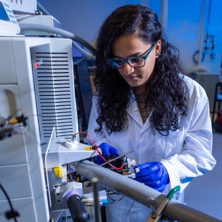 A scientist wearing a white lab coat, blue protective gloves, and glasses works intently with laboratory equipment. She has long dark curly hair and is focused on adjusting or repairing technical instrumentation with red and yellow wiring components. The image is set in a laboratory environment with a blue-tinted background and shows various scientific equipment and machinery. 