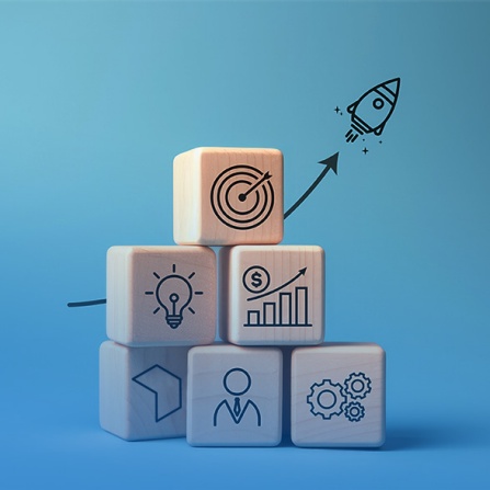wooden blocks stacked in a pyramid with symbols of commerce and business on thme and a rocket ship taking off in the background. 
