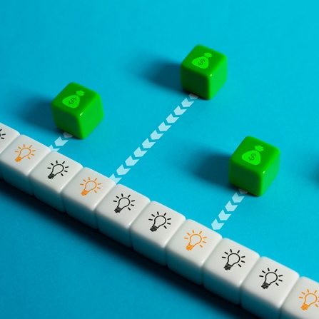 Green cubes with money bag icons move along arrow paths toward a row of white blocks printed with light bulb symbols, representing ideas turning into funding or investment opportunities. 
