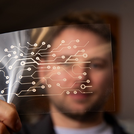A person holds a transparent panel printed with a copper circuit pattern in front of their face. 