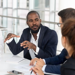 Three professionals sit at a table in a bright office space, with one person gesturing while speaking and the others listening, notebooks and a phone visible during a business discussion. 