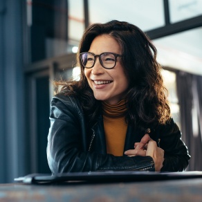 A smiling woman with dark hair and glasses sits at a table indoors, wearing a leather jacket and turtleneck, looking off to the side in a relaxed, conversational setting. 