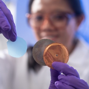 A person wearing protective gloves holds two circular lab samples—one translucent blue and one metallic—while standing in a laboratory environment, with their face softly out of focus in the background. 