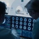 In Control Room Doctor and Radiologist Discuss Diagnosis while Watching Procedure and Monitors Showing Brain Scans Results, In the Background Patient Undergoes MRI or CT Scan Procedure. 