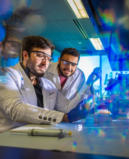 Two men in lab coats in a laboratory setting using test tubes and syringes. 