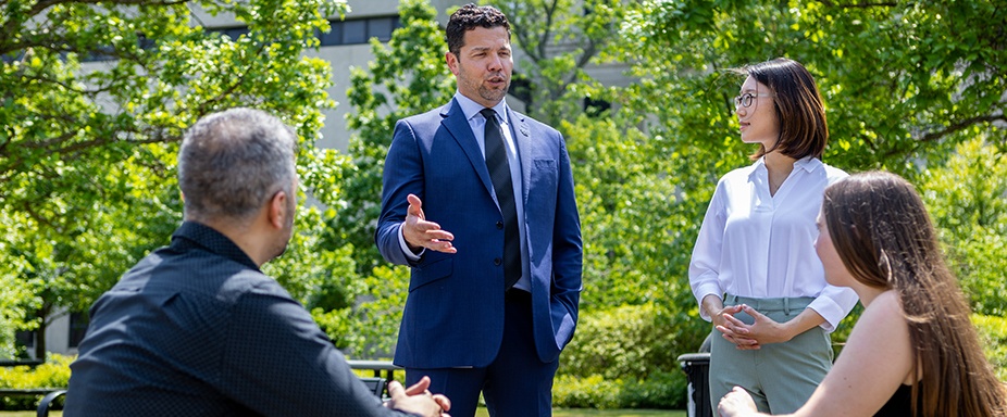 Group of university students listening as a suited advisor speaks with a student outdoors on campus.