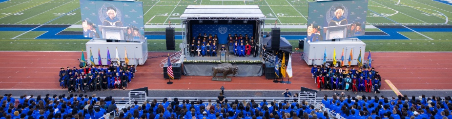 A large group of students wearing blue shirts sit in the stands. A stage and two screens are positioned on the track of Broadview Stadium. 