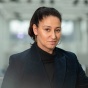 A woman in all black looks at the camera. Her hair is in a pony tail and she has hoop earings. 