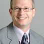 Photo of man in a grey suit with pink shirt and purple tie. He is smiling at the camera and has wire framed glasses. 