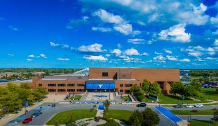 Aerial view of Alumni Arena at the University at Buffalo on a clear day, showing the main entrance, surrounding walkways, students, and campus greenery under a bright blue sky with scattered clouds. 