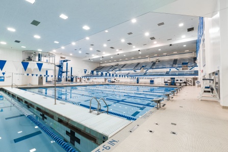 Wide view of the indoor competition swimming pool with multiple lap lanes, blue-and-white lane dividers, and stadium-style seating. Bright overhead lighting illuminates the natatorium, which features diving platforms, lifeguard chairs, and University at Buffalo branding throughout the aquatic facility.