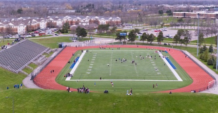 Aerial view of a large outdoor stadium with a green turf field surrounded by a red running track. Students and visitors are spread across the field, track, and bleachers during recreational activities on a cloudy day, with campus housing and parking lots visible in the background.
