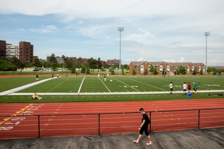Wide view of an outdoor athletic field with artificial turf and a surrounding red running track on a college campus. Students are walking, training, and relaxing around the field while campus buildings, trees, and tall stadium lights are visible under a partly cloudy sky.