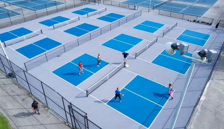 Aerial view of multiple outdoor pickleball courts with bright blue playing surfaces and gray surrounding pavement. Several people are actively playing doubles matches while others walk nearby. Black fencing surrounds the courts, and tall light poles overlook the recreation area on a sunny day.