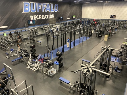 Elevated view of UB Recreation&rsquo;s fitness center, featuring weight machines, free weights, squat racks, and students exercising throughout the facility. 