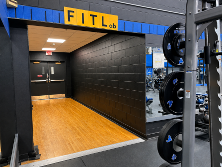 Entrance to the FITLab fitness space, featuring a yellow “FITLab” sign above a black-walled hallway with wood-look flooring, black double doors at the back, and a strength machine with weight plates visible on the right.
