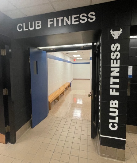 Entrance to Club Fitness, featuring black walls, bold white signage, and an open blue door leading to a brightly lit hallway. 