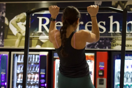 UB student participating in the pull-ups HardCORE Challenge, hanging from a pull-up bar inside Alumni Arena with vending machines in the background. 