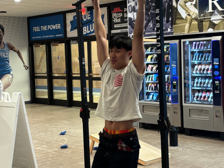UB student participating in the Dead Hang HardCORE Challenge, hanging from a pull-up bar inside Alumni Arena with vending machines and Buffalo athletics signage in the background. 