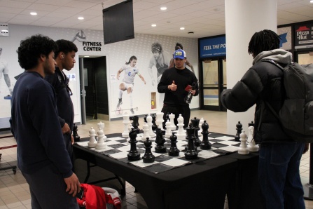Four students gather around a giant chessboard in a recreation center as one player gives a thumbs-up. 
