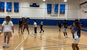 Students play a recreational basketball game on an indoor court in the Ed Wright Practice Facility at the University at Buffalo Recreation Center. Several players are spread across the court while a student staff member on the sideline drinks from a water bottle and observes the game. UB banners and a scoreboard hang above the court in the background. 