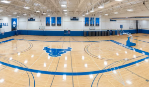 A wide view of the University at Buffalo Recreation Center basketball courts in the Ed Wright Practice Facility shows multiple empty hardwood courts with blue Bulls branding, adjustable basketball hoops, and championship banners hanging from the ceiling. The large UB Bulls logo is centered on the main court, with bright overhead lighting illuminating the spacious gym. 