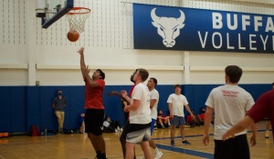A student attempts a layup during an indoor recreational basketball game at the University at Buffalo Recreation Center as several players watch the ball approach the basket. Other students wait along the sideline in the background beneath UB Bulls signage and gym banners. 