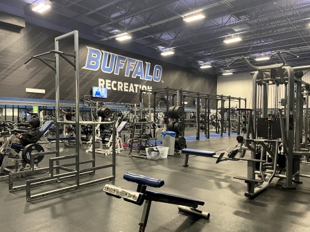 A spacious UB Recreation fitness center featuring weight machines, benches, squat racks, and free weights beneath a large &ldquo;Buffalo Recreation&rdquo; wall graphic. 