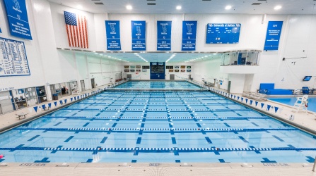 Wide view of the University at Buffalo indoor competition pool featuring multiple lap lanes with blue-and-white lane dividers, diving boards, championship banners, an American flag, a large scoreboard, and pool deck seating. The bright, empty natatorium is set up for swimming practice or competition.