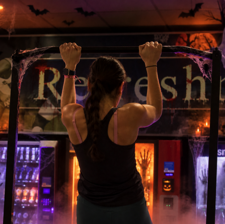 A person hangs from a pull-up bar in the foreground while a Halloween-themed gym scene glows behind them with animated-style pumpkins, bats, ghosts, cobwebs, and purple-orange lighting.