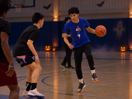 A student dribbles a basketball while facing a defender in an indoor gym, with other players nearby and a subtly dimmed Halloween backdrop featuring soft fog, glowing pumpkins, and faint ghost and bat projections on the wall.