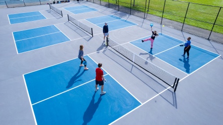 Aerial view of multiple outdoor pickleball courts with five people playing doubles on a sunny day. Players are spread across the blue and gray courts, holding paddles and actively engaged in a rally near the net, surrounded by black fencing and green grass.