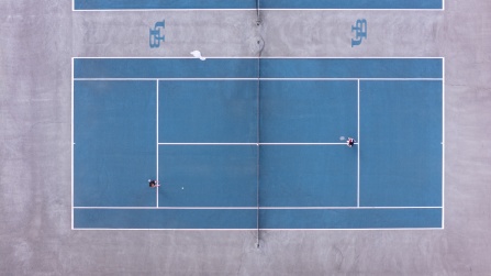Aerial view of two people playing tennis on a blue outdoor court at the Ellicott Tennis Complex on UB North Campus. The image captures the court from directly above, showing the players positioned on opposite sides of the net during a rally.
