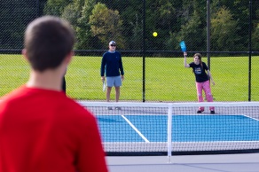 A young woman returns a bright yellow ball during an outdoor pickleball game as her teacher and an opposing player watch.
